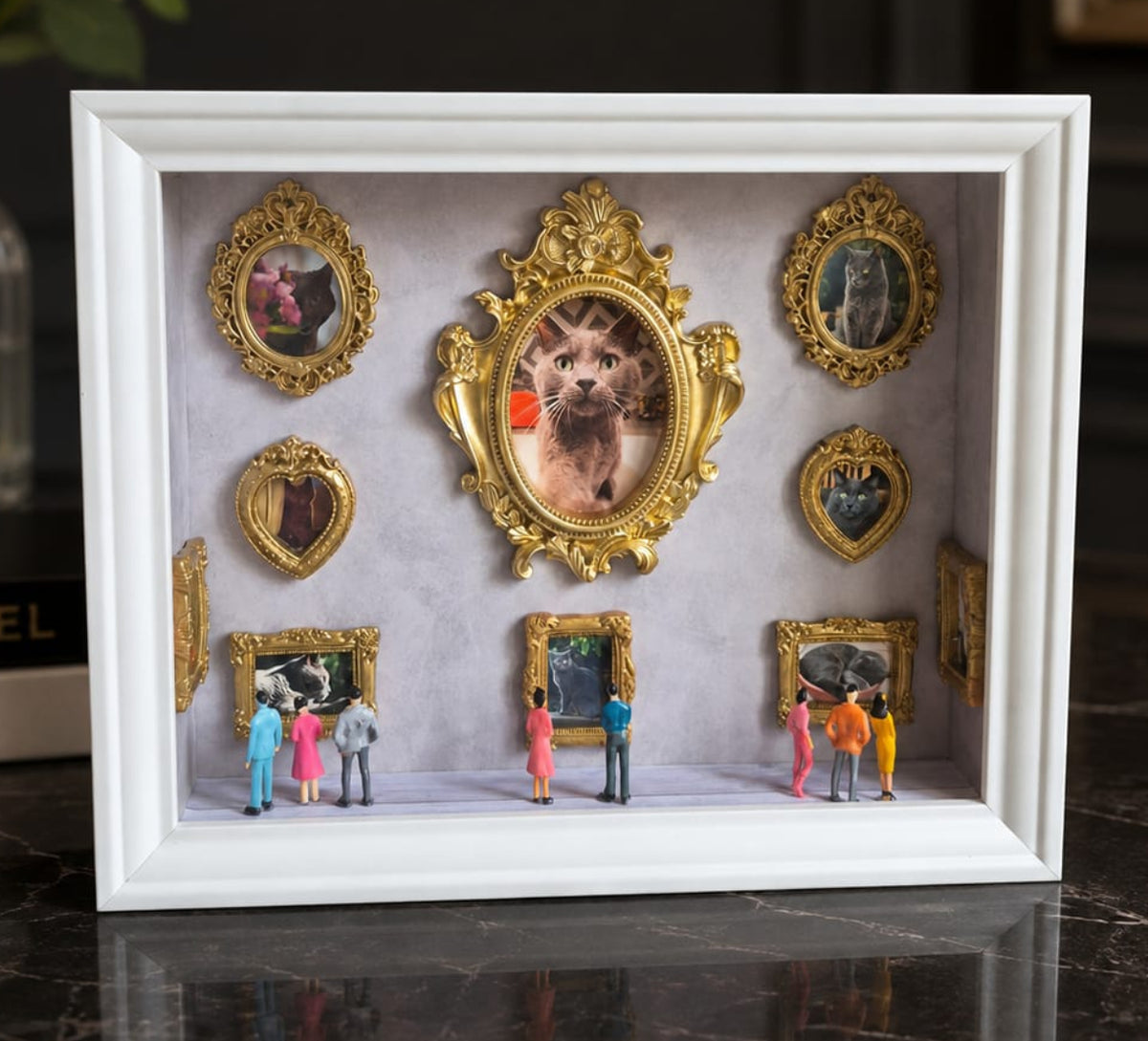 A white shadow box display frame with ornate gold  mini frames and miniature figurines, featuring a pet portrait in the center, styled on a dark marble table, museum of us. Pet pictures