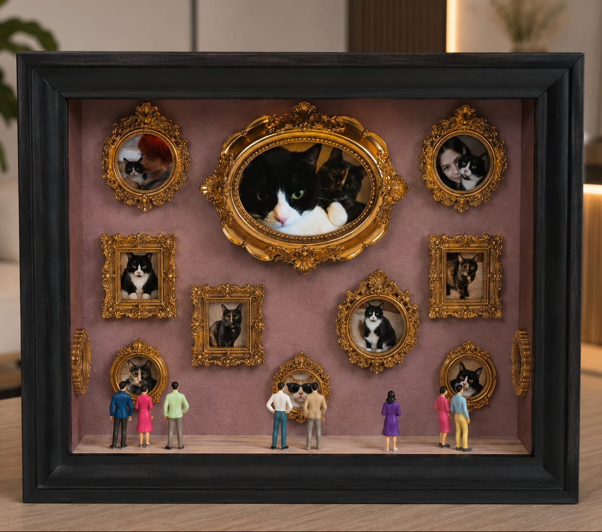 A framed shadow box display on a tabletop, featuring multiple ornate bronze mini frames with pets portraits arranged on a dusty pink background, with tiny figurines standing in front. The outer frame is black, and the scene is photographed in a real indoor setting with blurred furniture and decor in the background.