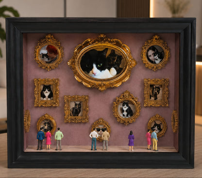A framed shadow box display on a tabletop, featuring multiple ornate bronze mini frames with pets portraits arranged on a dusty pink background, with tiny figurines standing in front. The outer frame is black, and the scene is photographed in a real indoor setting with blurred furniture and decor in the background.